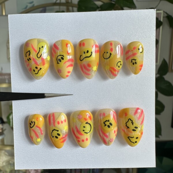 Neon swirls of pink and orange with smiley face decals and star accents on a golden yellow base. The design features fiery orange, hot pink, bold black, and pops of pastel with a glossy, vibrant finish.