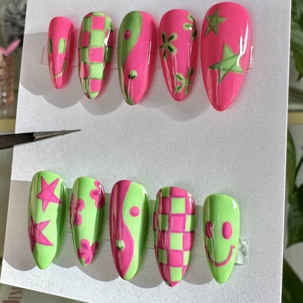Mismatched hand painted nail art featuring 3D smiley faces, yin-yang symbols, checkerboards, groovy flowers, and bold starbursts. The design incorporates neon pink and electric lime green with a glossy finish.