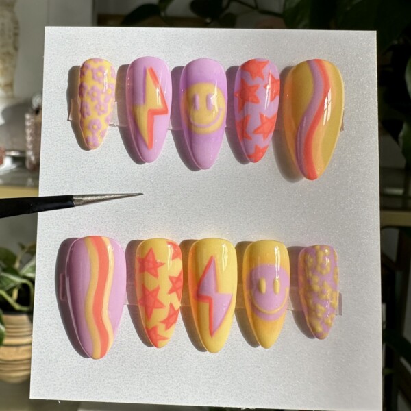 Mismatched hand-painted nail art featuring retro smiley faces, power bolts, daisies, shooting stars, and mod-inspired wave patterns. The design incorporates lavender, lemon yellow, neon pink, and warm orange with a glossy, buttery smooth finish.