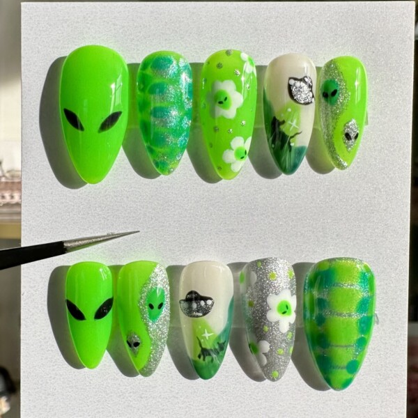 Custom press-on nail set featuring hand-painted alien and space-themed designs. Bright purple or neon green tones, glitter accents, florals, and extraterrestrial art on a short almond shape. Glossy finish and reusable nails for bold, statement-making looks.