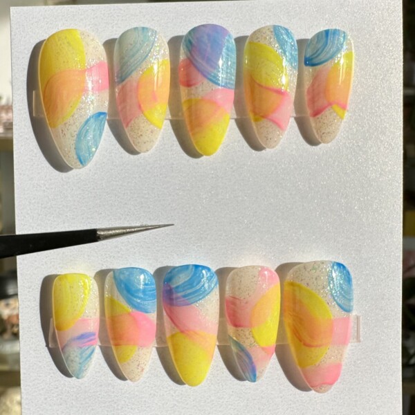 Custom press-on nail set featuring hand-painted abstract designs inspired by primary colors. Bright pink, blue and yellow swirls and blobs layered over a soft nude base and a hint of glitter on a medium almond shape. Glossy finish and reusable nails for bold, statement-making looks.