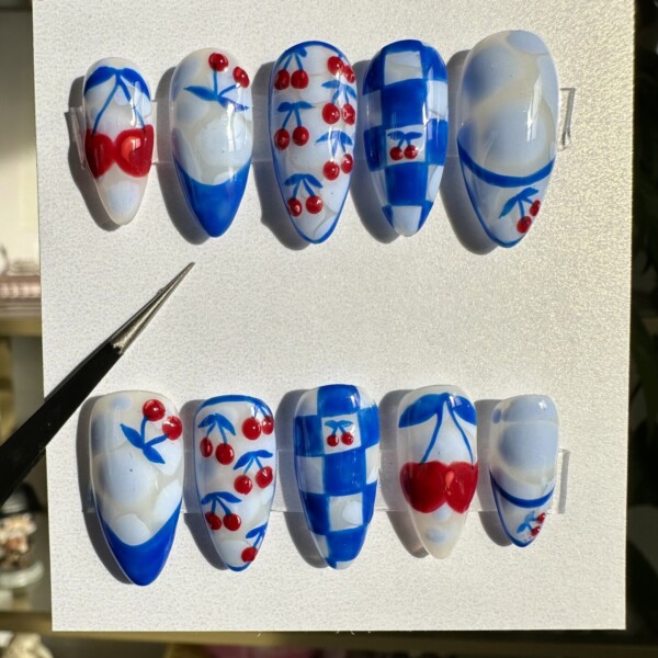 Hand-painted press-on nails with mismatched cherry art, cobalt blue checkerboard patterns, soft cloud-like swirls, and bold red accents. Glossy, high-shine finish on a retro-inspired design. Fun, fruity, and sky-themed nail set.