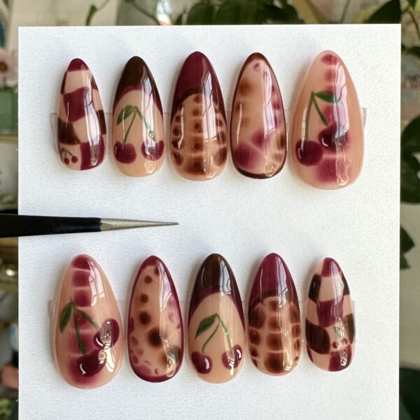 Hand-painted press-on nails in deep cherry red, blush, and nude tones with cherries, checkerboard patterns, gradients, and dot details. Medium almond shape with a glossy finish. Bold, artsy, and fruity nail design with a moody twist.