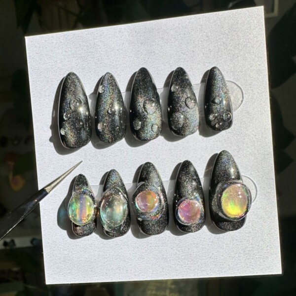 A set of medium almond press-on nails with a glossy black base and holographic glitter, resembling a starry night sky. Five nails feature iridescent, bubble-like domes that reflect rainbow hues. The design gives off a mysterious, celestial vibe with a futuristic edge.