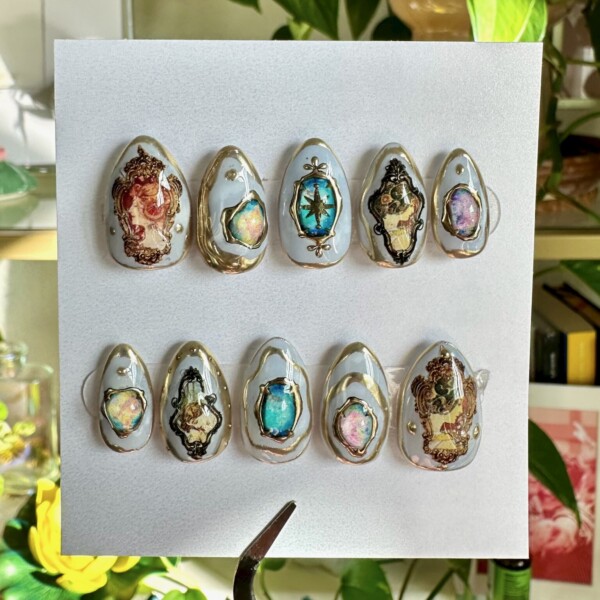 A luxurious set of ten press-on nails in a short almond shape, painted with a soft milky white base. Each nail features ornate gold borders framing detailed baroque-style cameos, iridescent gemstone accents, and romantic mythological illustrations. The design resembles miniature vintage portraits, evoking an old-world, gallery-like elegance. Set is photographed against a sky-blue background, enhancing its dreamy, regal aesthetic. Ideal for fans of art history, opulence, and storytelling through nail design.