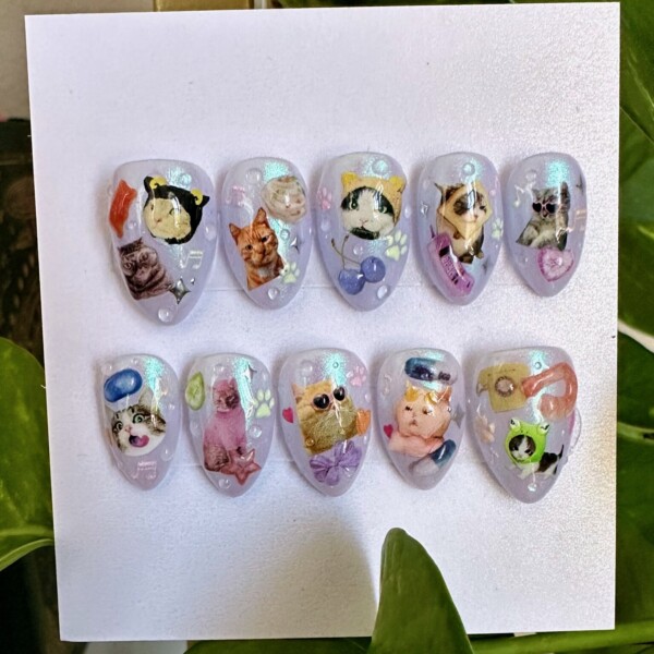 Short almond press-on nails with an iridescent shimmer base featuring playful illustrated cats in various quirky poses and accessories, including sunglasses, hats, bows, and fruit, plus tiny 3D embellishments like hearts, paw prints, and musical notes.
