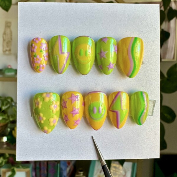 Mismatched hand-painted nail art featuring retro smiley faces, power bolts, daisies, shooting stars, and mod-inspired wave patterns. The design incorporates lemon yellow, neon green, and pale lilac with a glossy, buttery smooth finish.