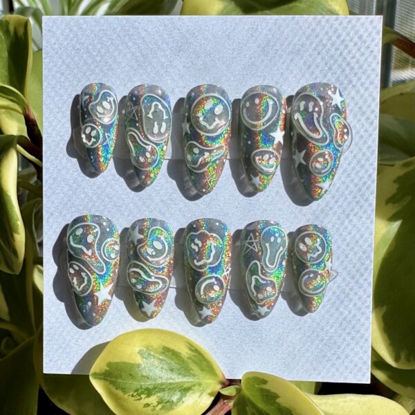 White chrome holographic press-on nails with playful white smiley faces, stars, and abstract squiggles in a medium almond shape, glowing under natural light.